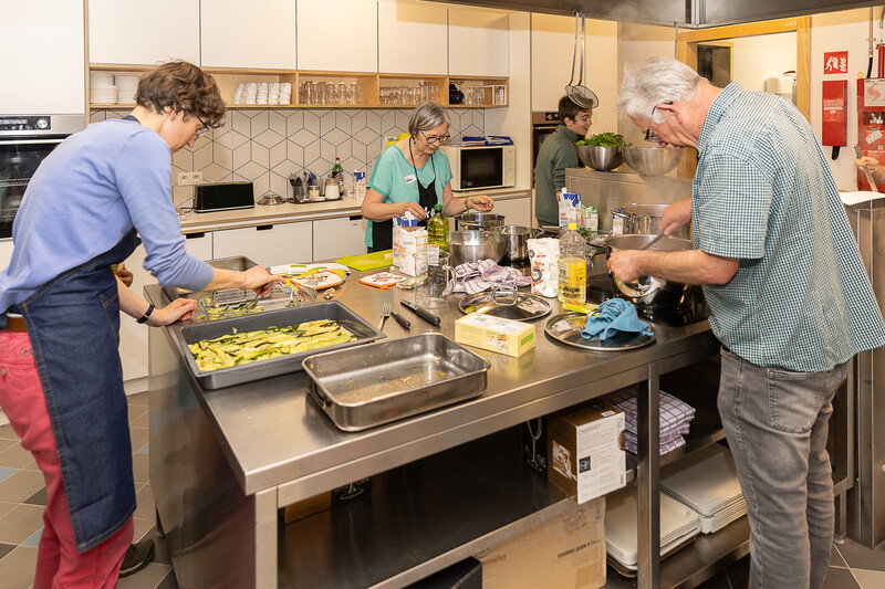 Vier personen werken aan een groot, inox werkblad in een semiprofessionele keuken om een gezonde maaltijd voor te bereiden. Een grote schaal courgette wordt afgekruid en iemand roert in een kookpot.