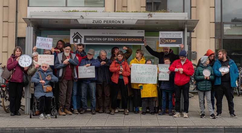 Kijkend op hun uurwerk en op klokken wacht een groep mensen ludiek in een bushokje op een rechtvaardig woonbeleid.