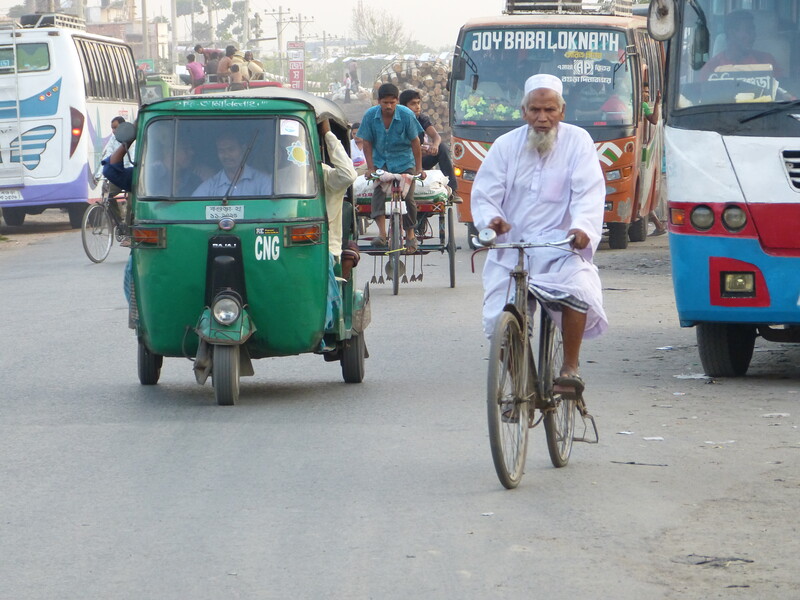 Oudere man in wit gewaad fietst in het drukke verkeer tussen bussen en tuktuks te Bangladesh.