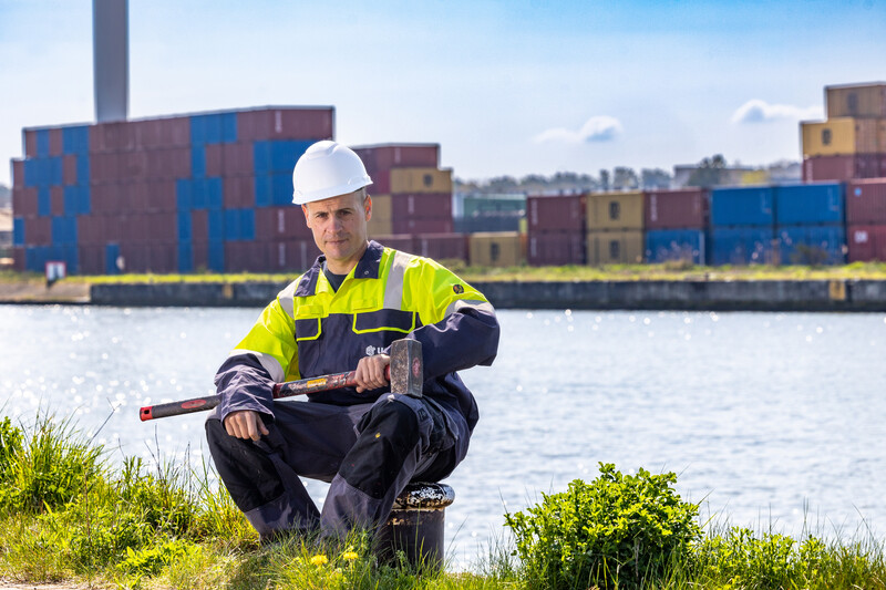 Een onderhoudsmecanicien zit met een hamer aan de rivier voor een industrieterrein