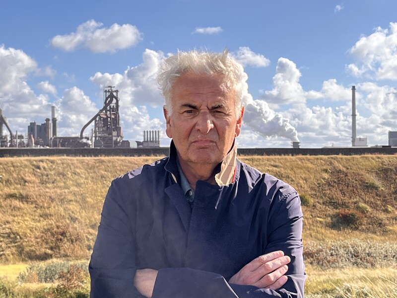 Een man van middelbare leeftijd poseert met gekruiste armen voor een landschap met in de achtergrond zware industrie.