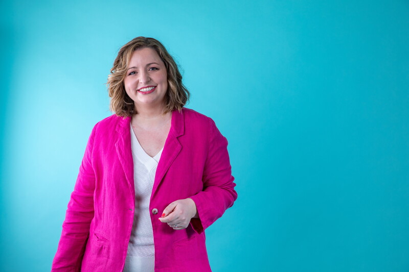 We zien een vrouw, Jacotte Brokken, met een felle roze vest op een blauwe achtergrond