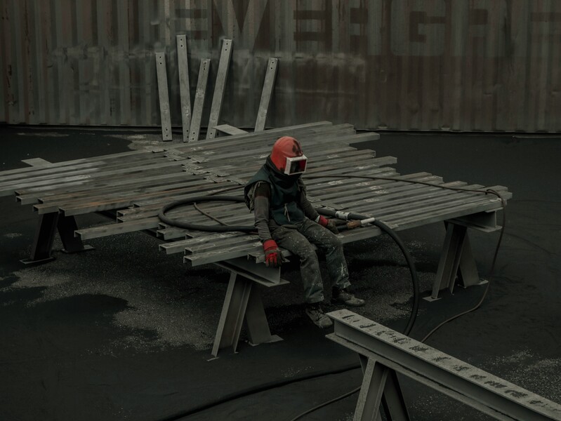 Een werker in beschermende kledij en zandstraalmateriaal staart besluitloos voor zich uit