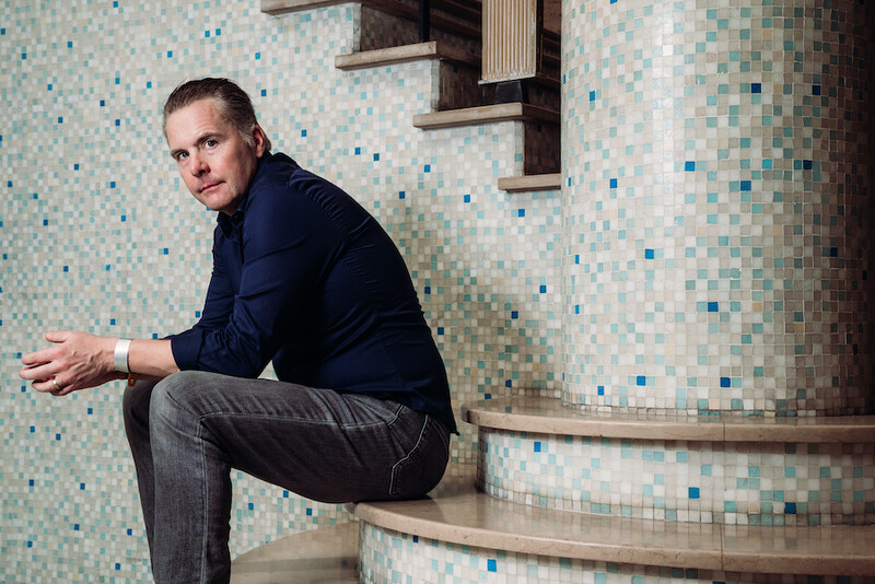 Bruno Fierens, het enige Belgische lid van Millionaires for Humanity, in hemd en jeans, gefotografeerd in zijaanzicht, zittend op treden voor een mozaïekmuur